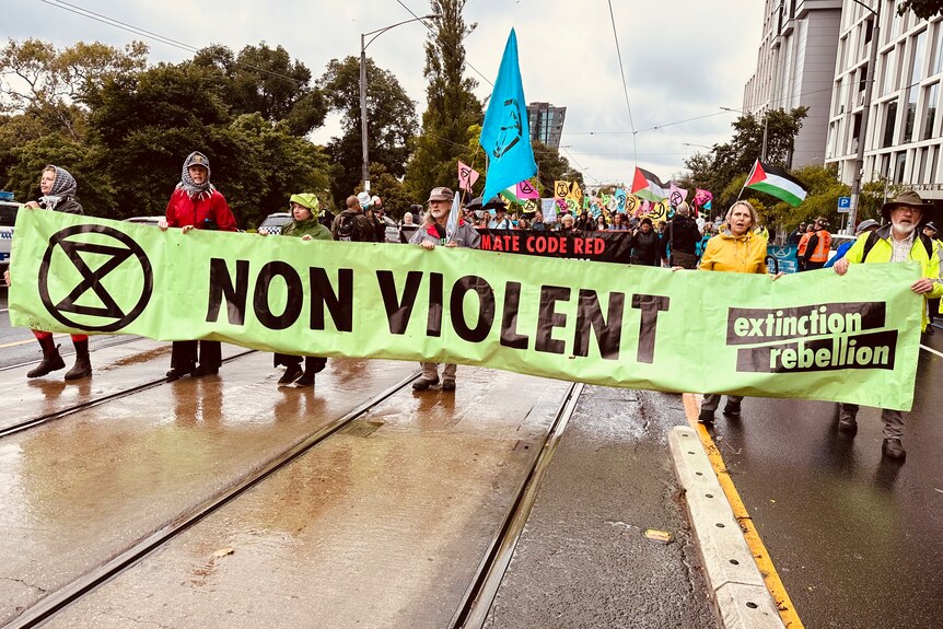 Six climate activists lead a march on a Melbourne road holding a long green Extinction Rebellion banner reading 'Non Violent'.