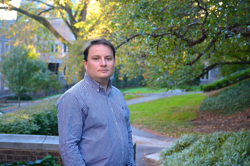 Man stands in front of a garden and trees with a blue shirt on, staring at camera. 