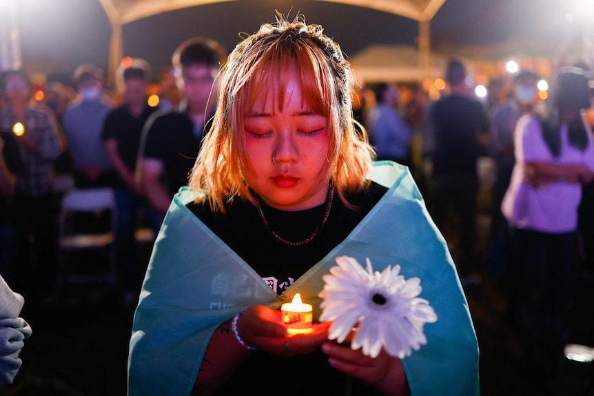 A person holds a candle and flower, covered in a blue cloth looking sombre