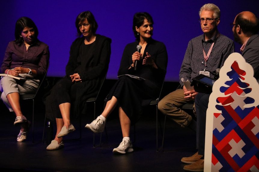 three women and two men sit on a stage. the woman in the center has a microphone and is talking