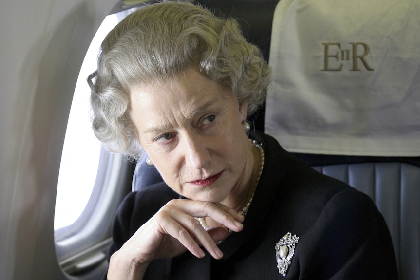 Helen Mirren, as the Queen, rests her head on her hand as she sits in a private jet and looks seriously to the right