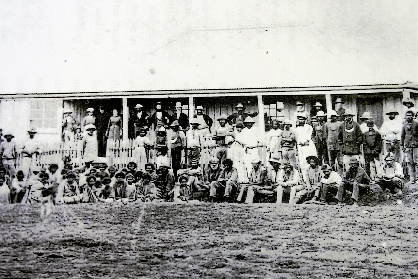 A black and white photo of a large group of Indigenous people standing and sitting near a building.