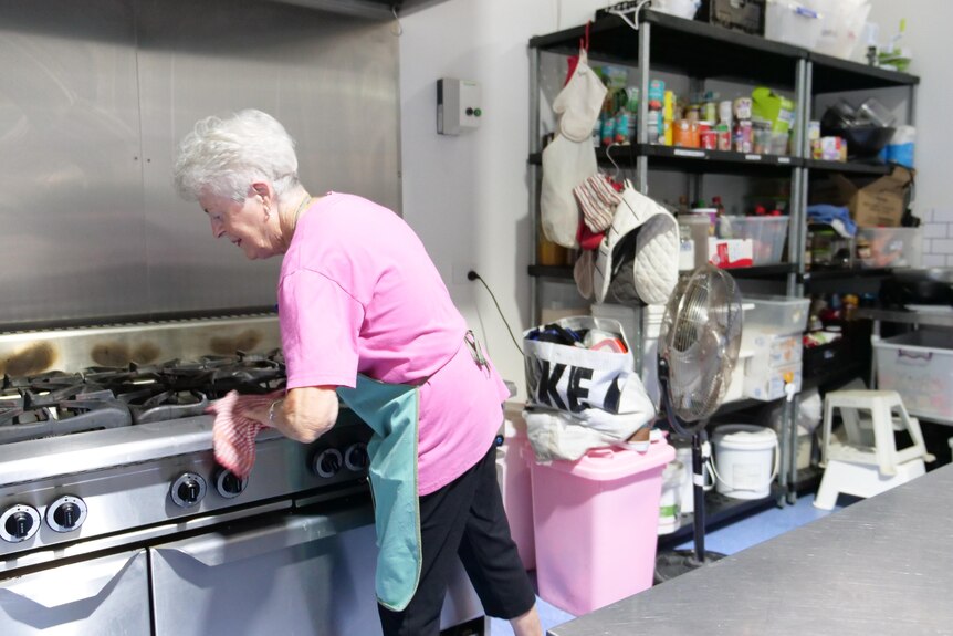 A senior woman leans over a large steel stovetop, cleaning it with a cloth. A large shelf of groceries is in the background.
