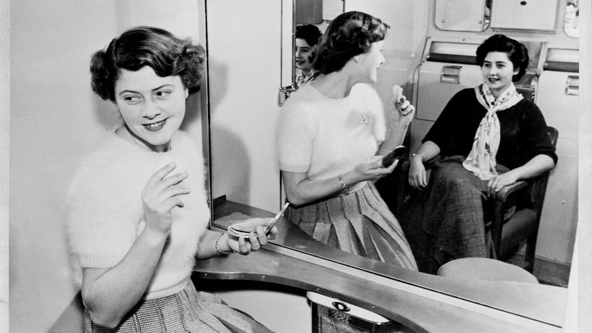 Two young women talk and apply make-up in a mirrored railway carriage.