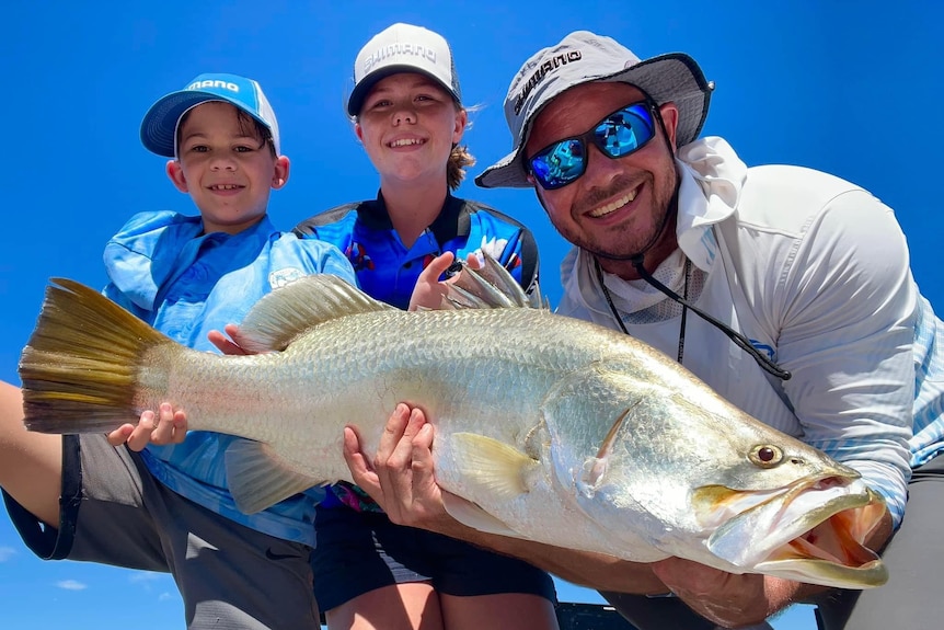 Two young children with a man holding a barramundi fish.