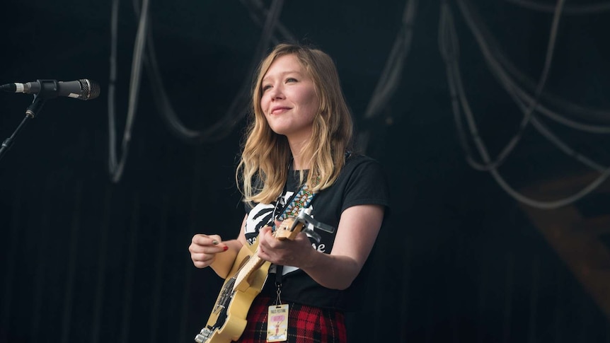 Julia Jacklin smiles while playing guitar live at Falls Festival against a black backdrop