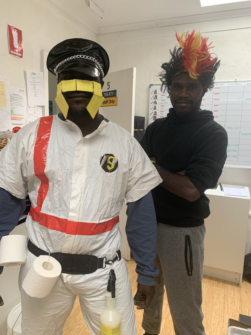 Two men dressed up stare at the camera, one is wearing a white suit with tape over his face and a hat.