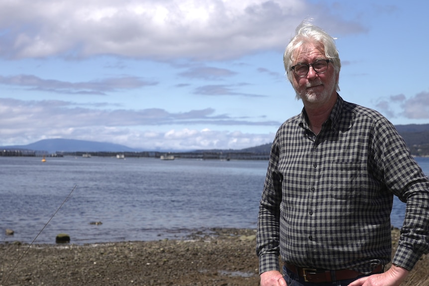 Man with white hair, wearing checked shirt, stands with his hands on his hips in front of an ocean area.
