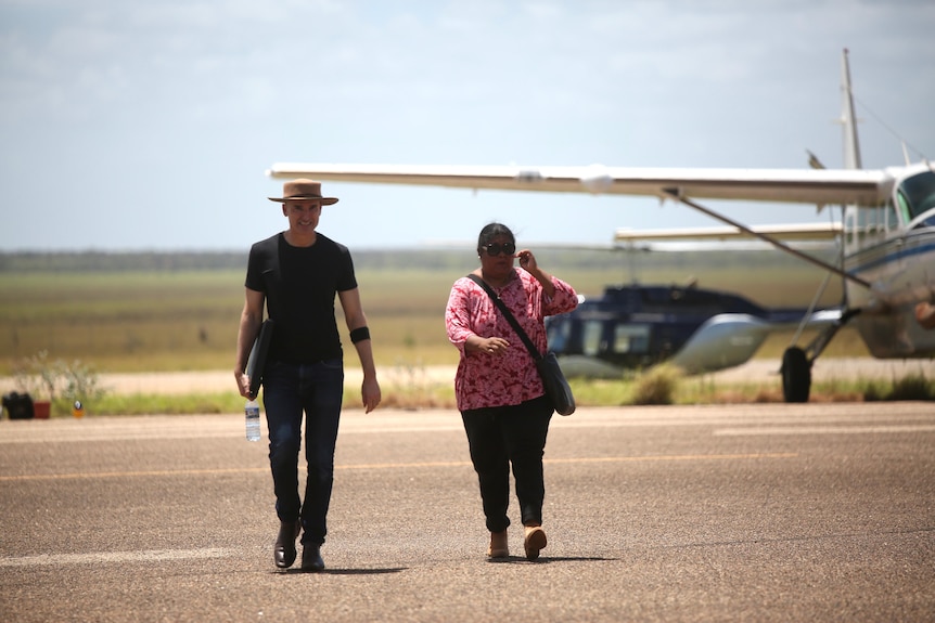 A man and woman crossing an airstrip with a plane in the background