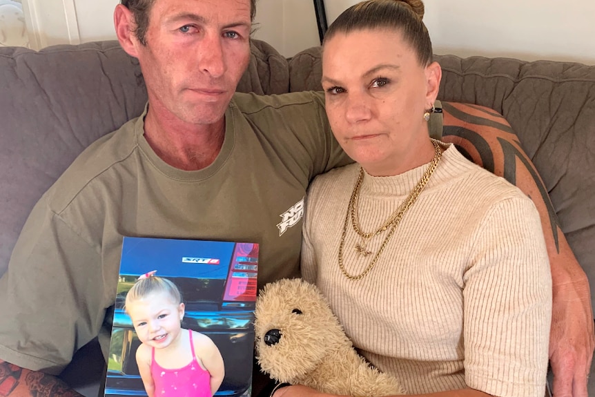 A husband and wife sit close on the couch, holding hands and looking sad while holding a photo of their toddler daughter