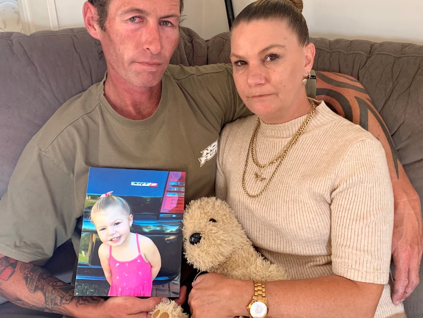 A husband and wife sit close on the couch, holding hands and looking sad while holding a photo of their toddler daughter