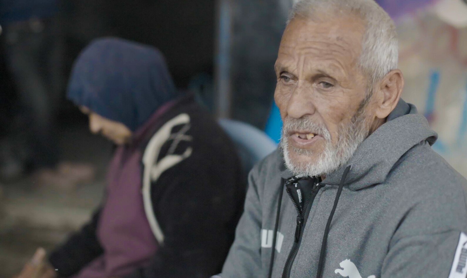 A man wearing a grey jumper sits next to a woman wearing a hijab as they talk.