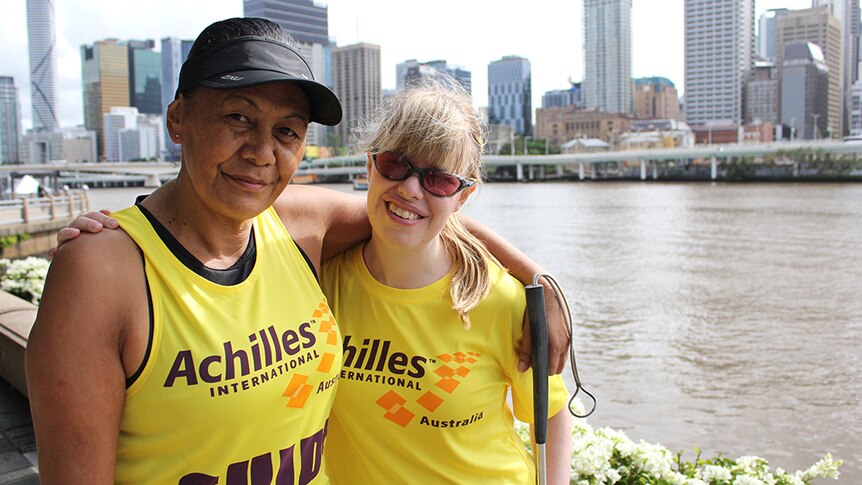 Two women in yellow shirts standing arm in arm in front of the Brisbane River.