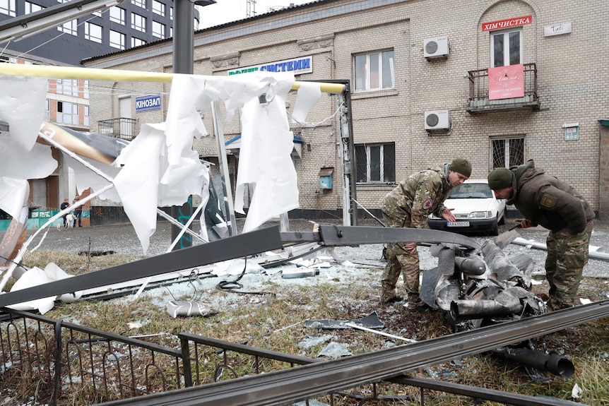 Two men dressed in military fatigues inspect a twisted mess of metal on the ground