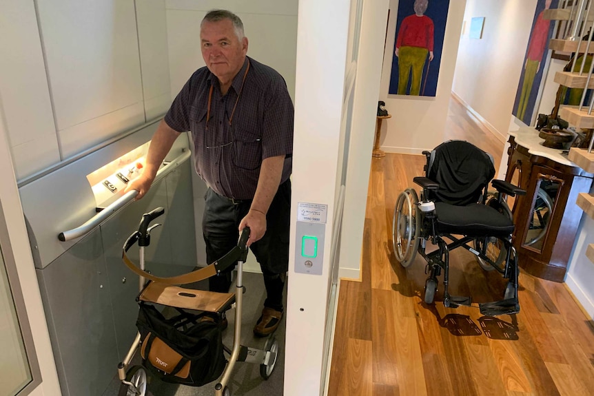 Peter Willcocks walks out of a lift in his home using a walker with his wheelchair nearby.