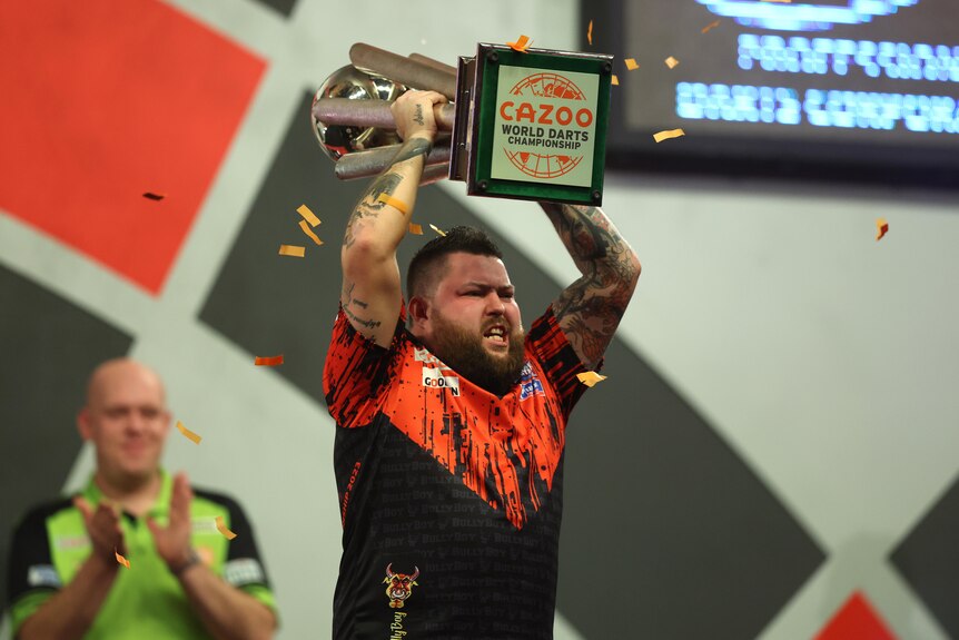 A darts player brandishes the world championship trophy above his head as confetti falls from the ceiling.  