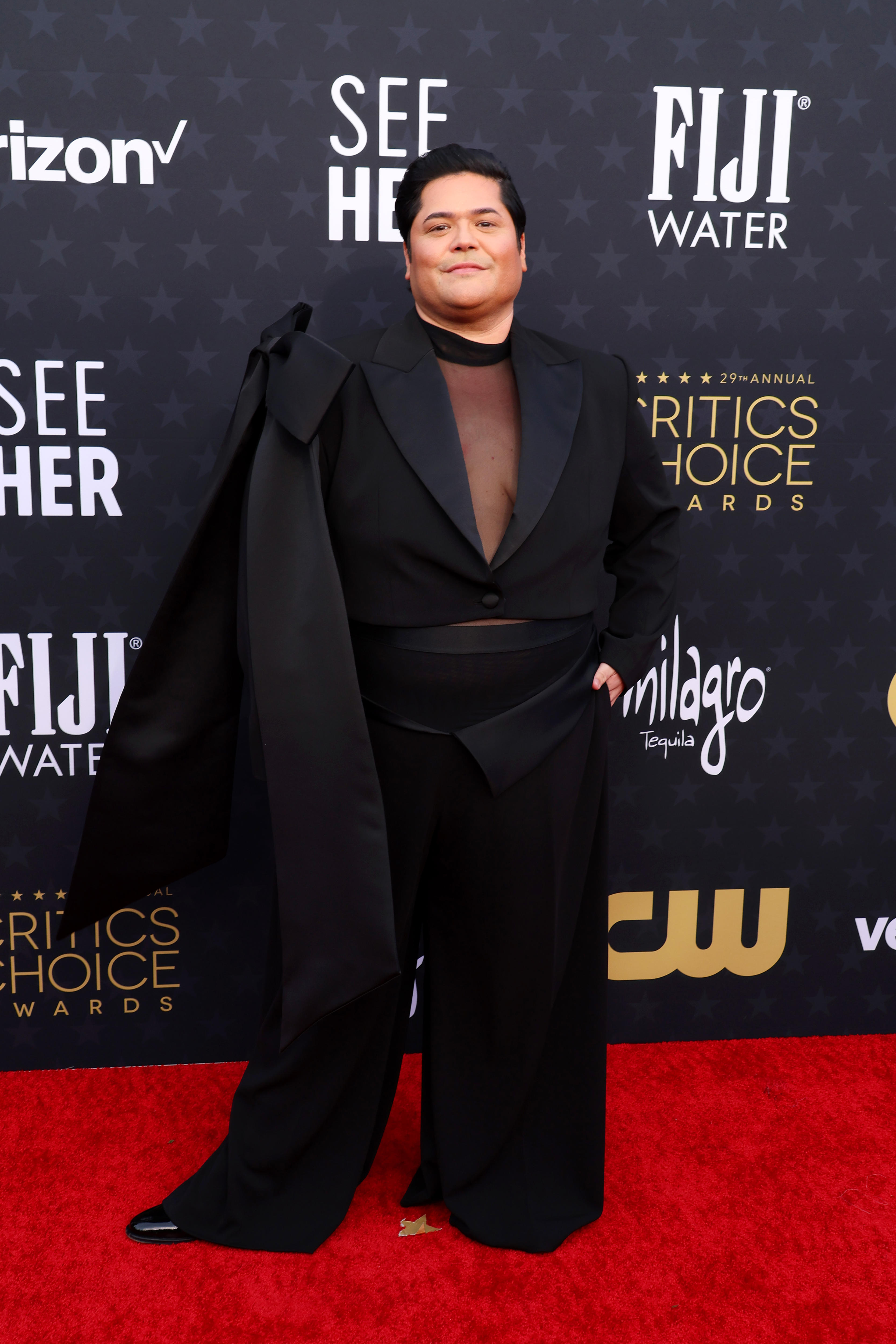 Harvey Guillen wearing a silky black suit with a sheer top underneath and a big bow on on of the sleeves