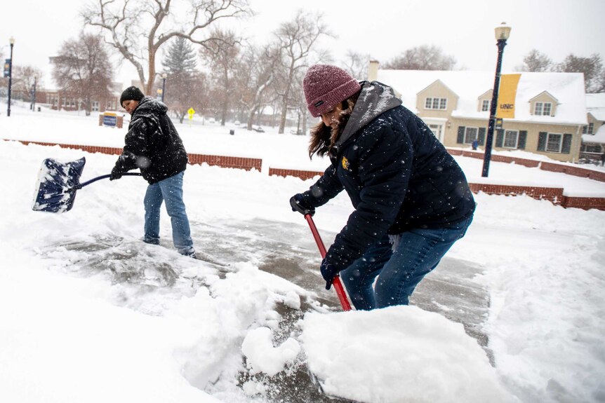 A man and a woman dressed in thick winter clothing shovel heavy snow from the steps of a building.