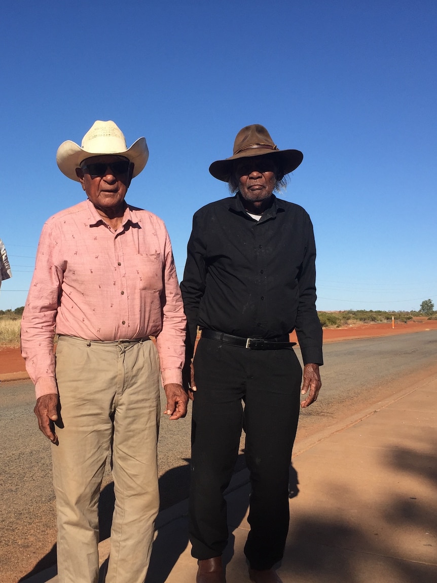 Two men wearing stockmen's hats and long-sleeved button-down shirts stand beside a rural road.