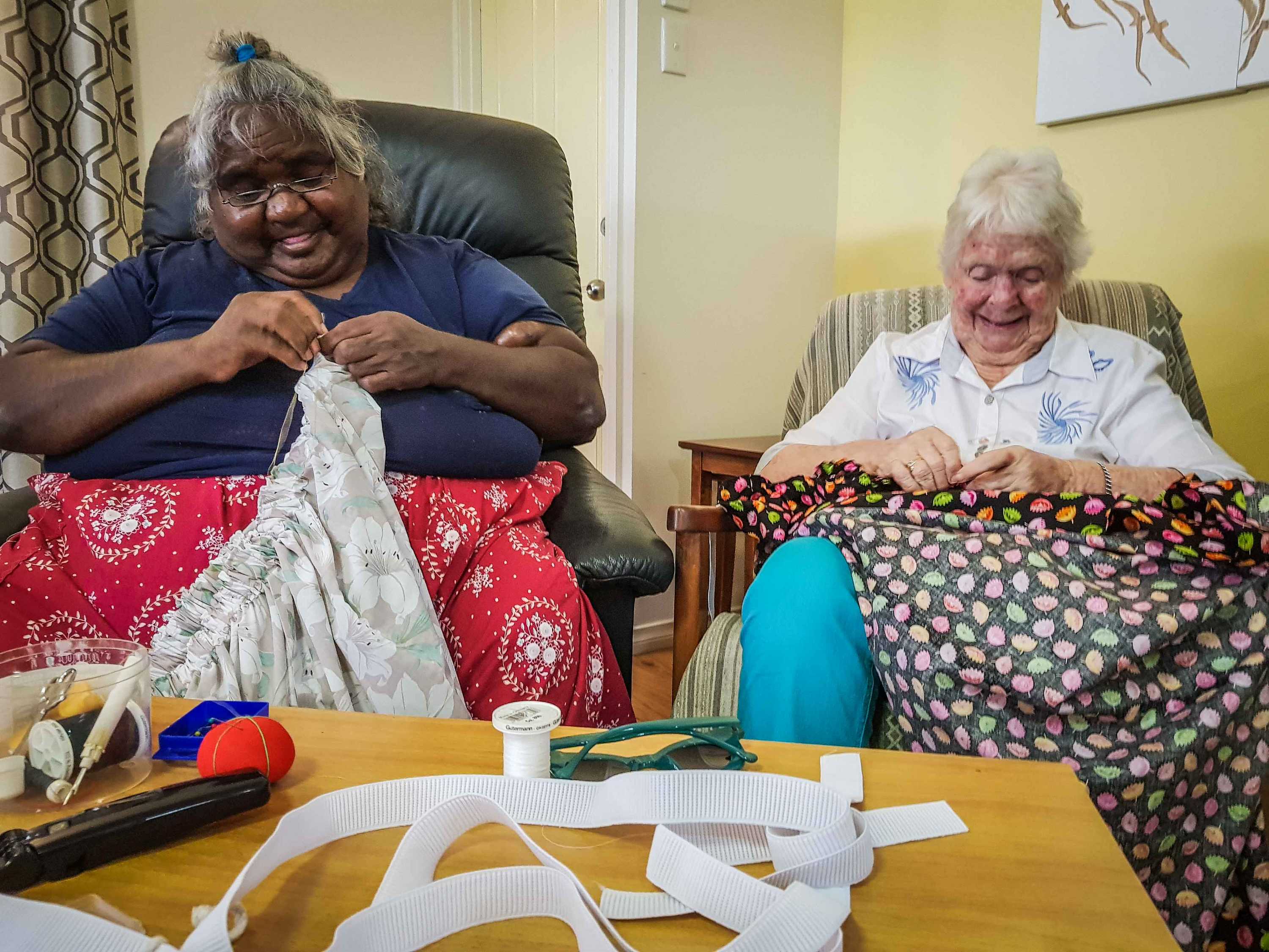 Two women sit in armchairs hand-sewing skirts.