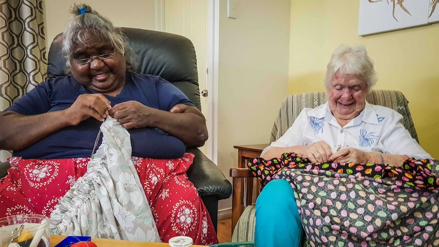 Two women sit in armchairs hand-sewing skirts.