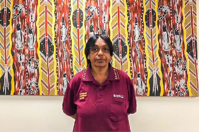 A woman is a corporate shirt stands in front of a large Indigenous arty work.