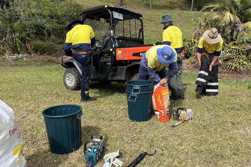 Four biosecurity officers are working on a grassy landscape outside, with hats on. They are working with tools and buckets.  
