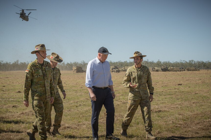 A man in business clothes walks with soldiers along a field, with a military helicopter in the distance.
