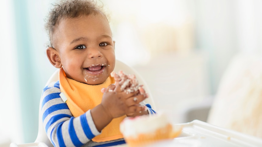 A baby feeds himself in a high chair while smiling.