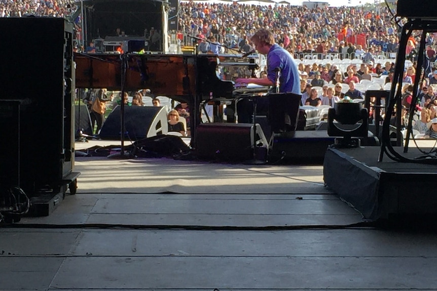 A man in a blue shirt playing piano in front of a large stadium crowd.