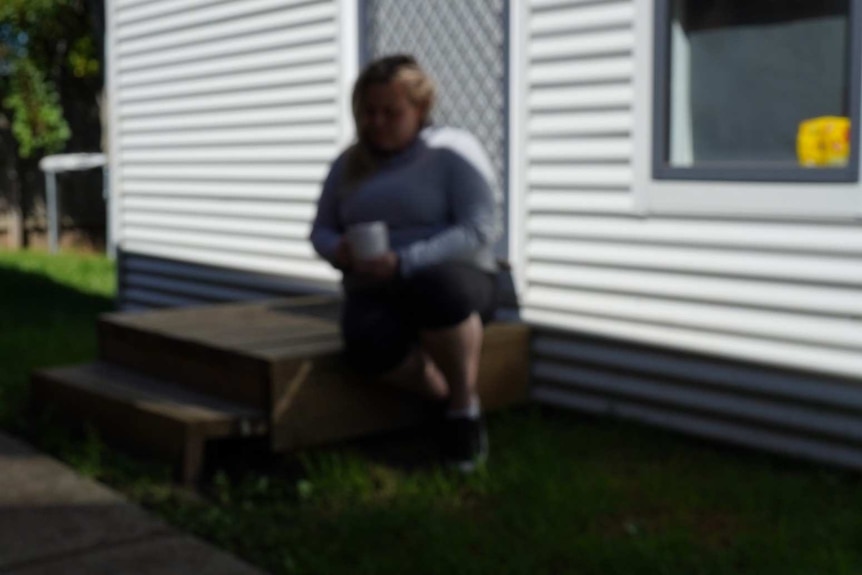 A woman is shown sitting on the step of a house, holding a cup.