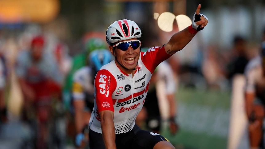A smiling Caleb Ewan raises one finger after crossing the finish line.