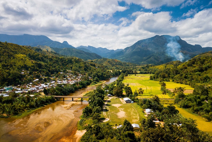Aerial village, river and green fields with mountains covered in dense jungle in background.