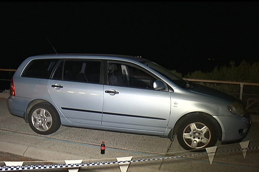 A small blue station wagon parked on the side of the road at night with police tape around it.