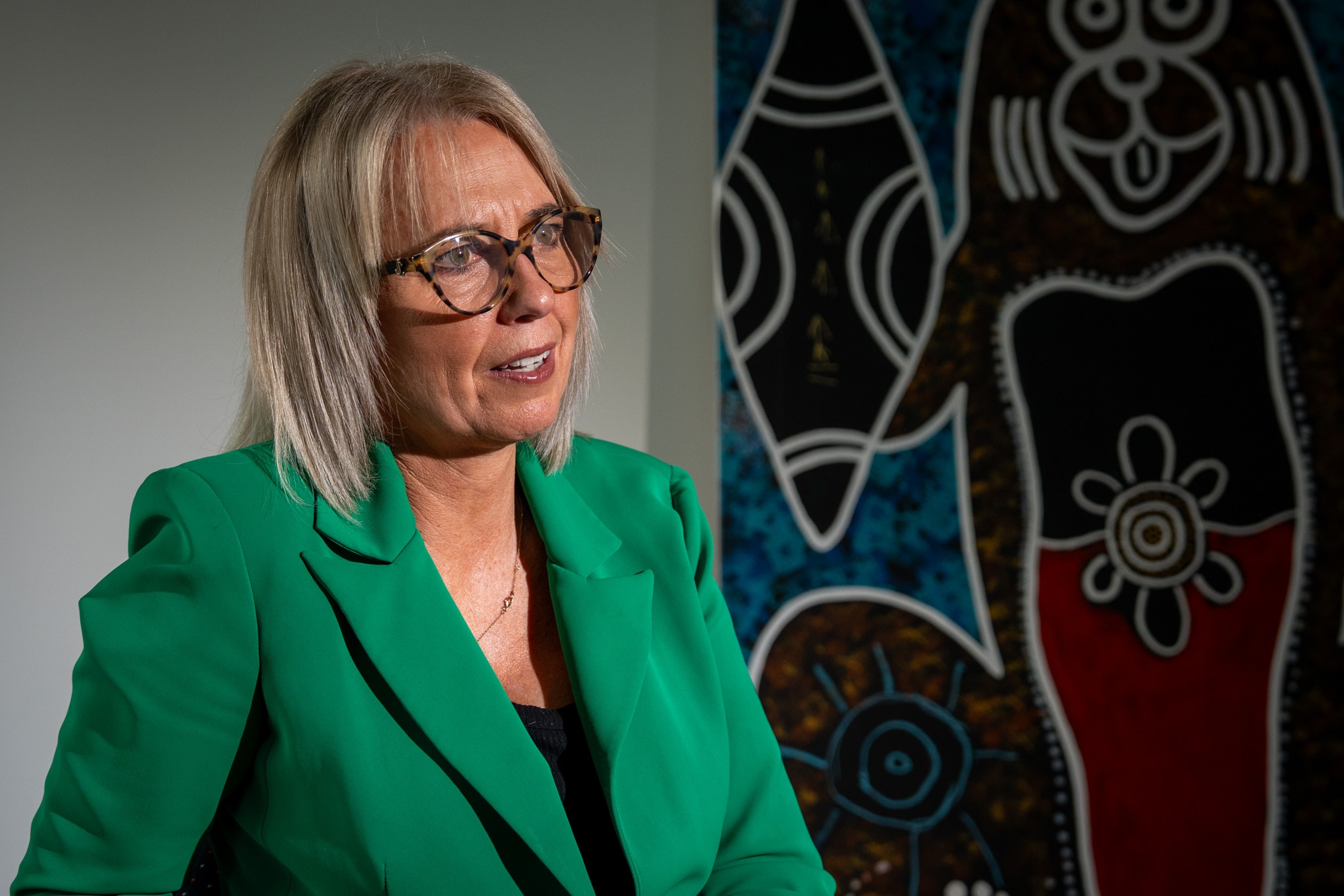 Woman wearing green blazer sitting in front of an Indigenous artwork.