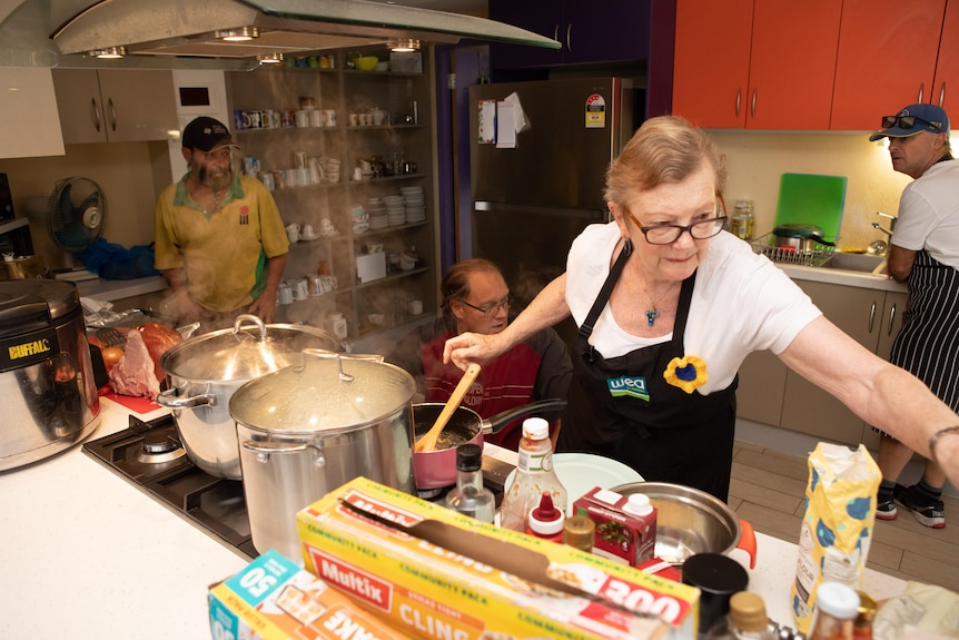 Olwyn-Ann busily cooks ina kitchen, others from the community gathered around her.