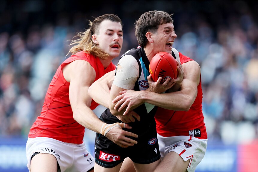 Zak Butters carries the ball while being wrapped up by two Essendon players