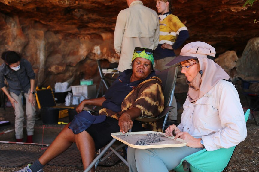 An Indigenous woman sits next to a white woman to sift through rocks in a cave. 