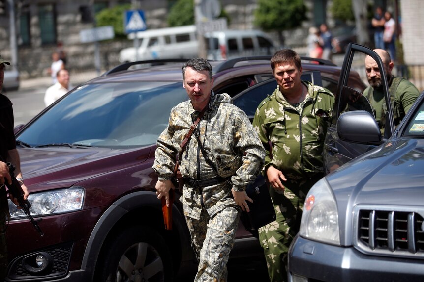 Two men dressed in military fatigues and holding guns walk past cars.