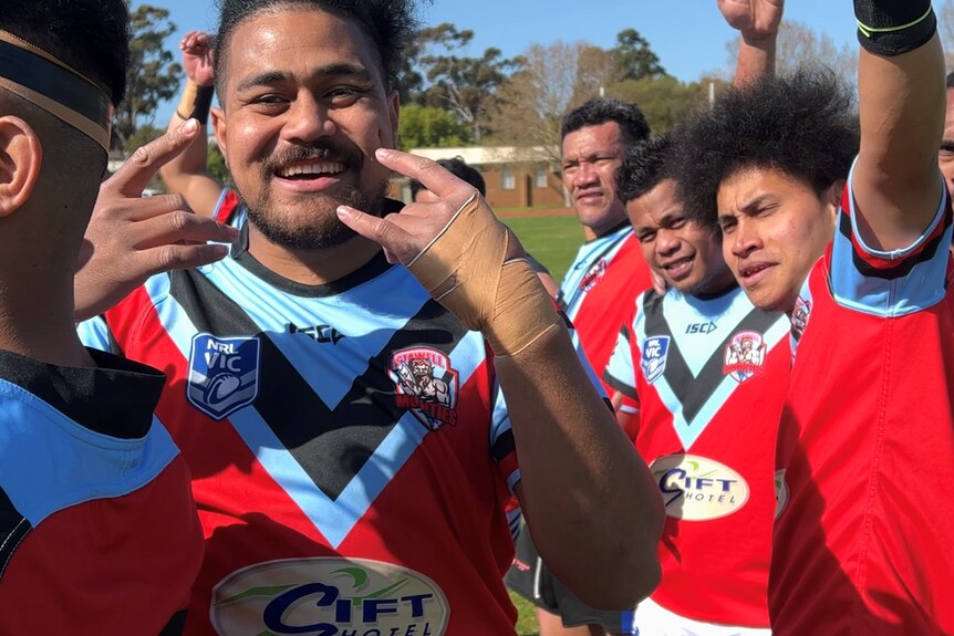 man wearing blue and red rugby jersey makes rock on gestures surrounded by team mates with fist pumps.