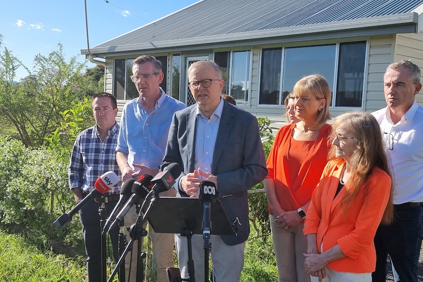 The prime minister wears an open light shirt, grey jacket, speaks into media mics, NSW premier,  group stands next to him.