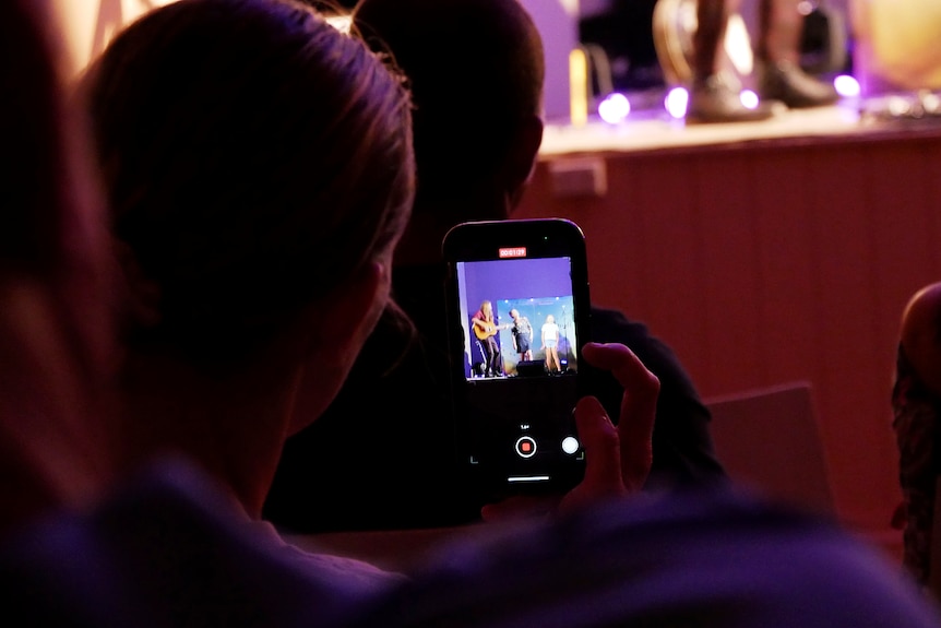 An audience member uses their phone to film three performers on a stage