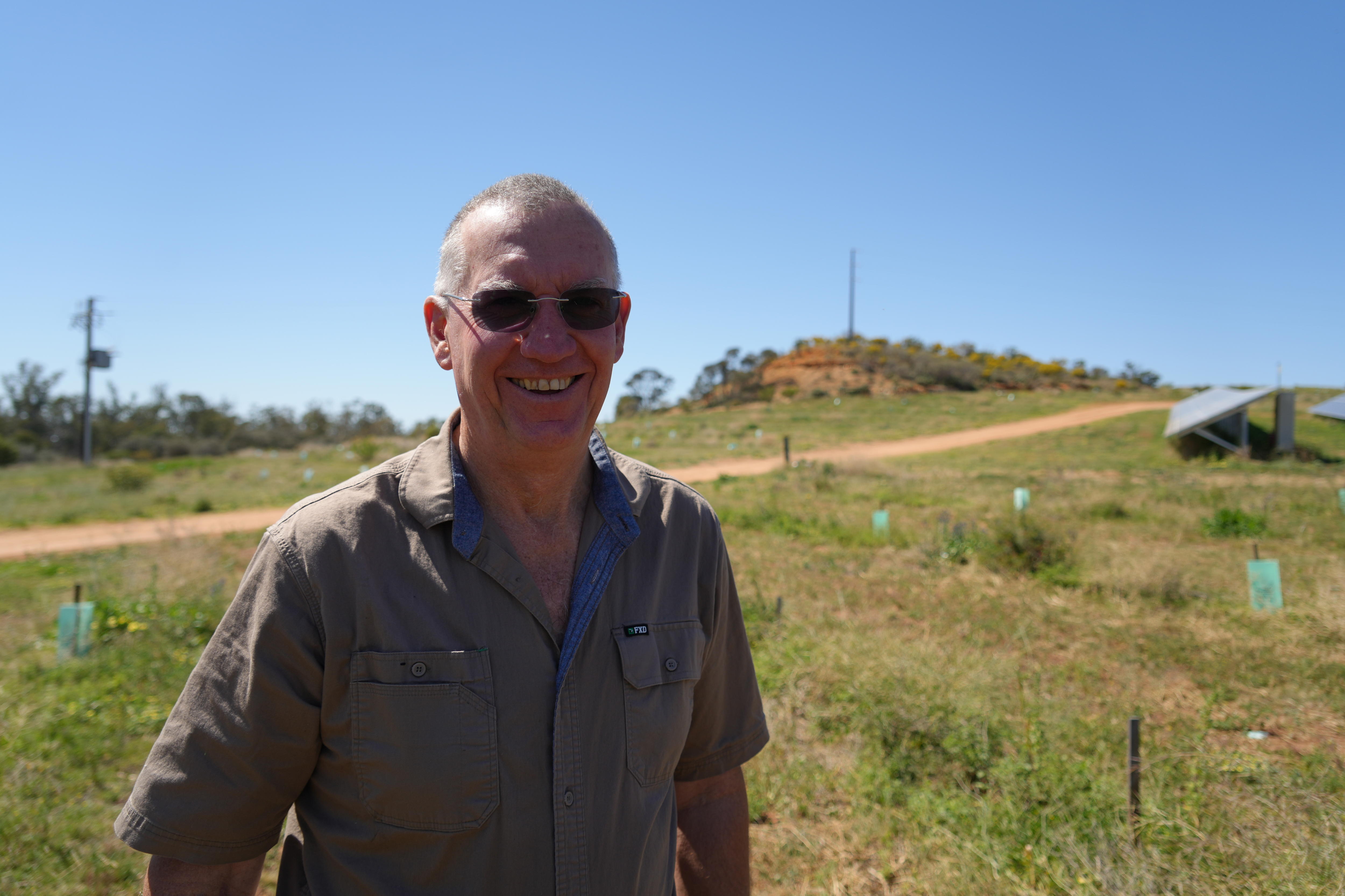 A man wearing sunglasses smiles while standing in front of native Australian vegetation.