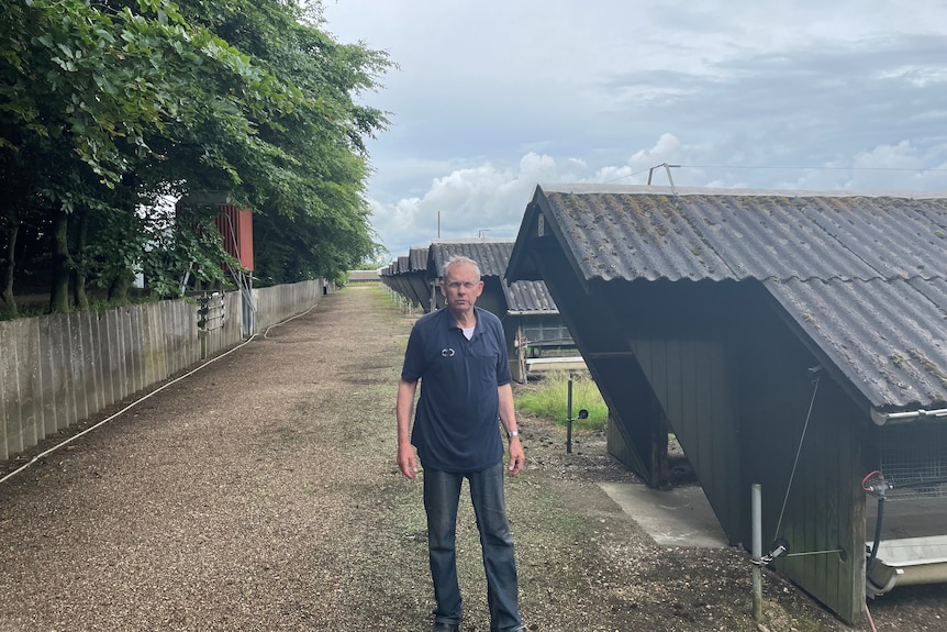 A middle aged man stands next to sheds looking stoic.