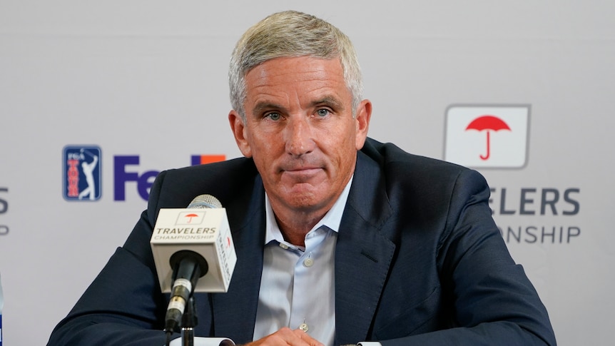 A top golf executive sits at a desk in front of a microphone and looks out at reporters at a press conference.