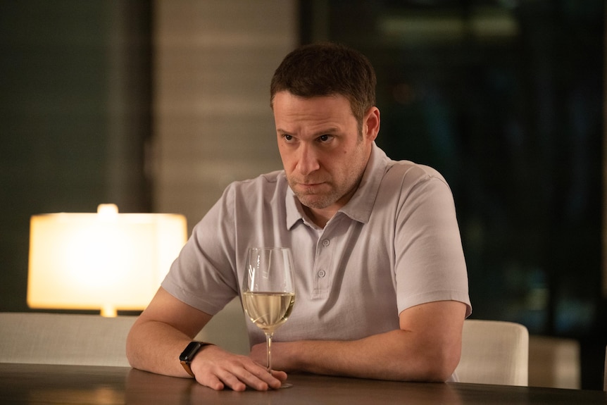 Middle-aged white man with short brown hair wears a cream polo shirt and sits at a kitchen bench drinking white wine.