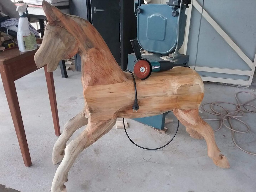 A rocking horse that has just been carved and glued together and sits in a shed with an electric sander perched on its back.