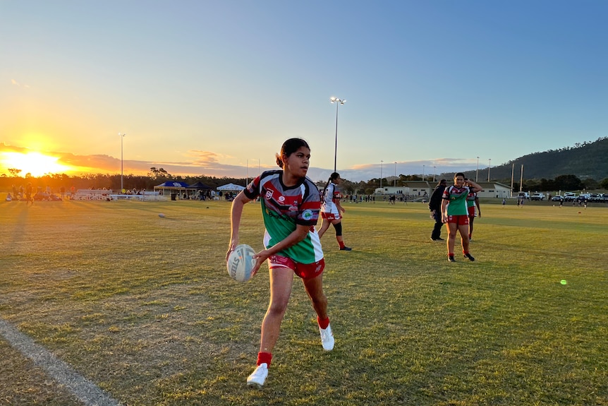 Lily passes the footy onto her team-mates on the field as the sun sets behind her. 