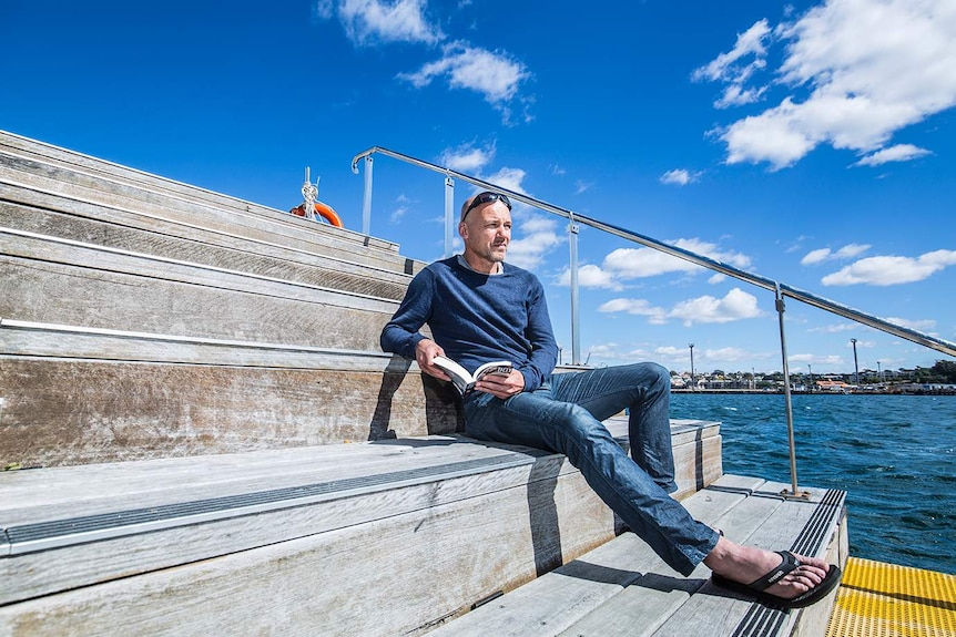 Colour photograph of Gary Jubelin reading a book in the sun by the water