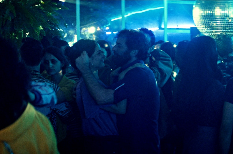 A woman and man hold each other closely on busy dancefloor in blue hue lit nightclub with disco ball.